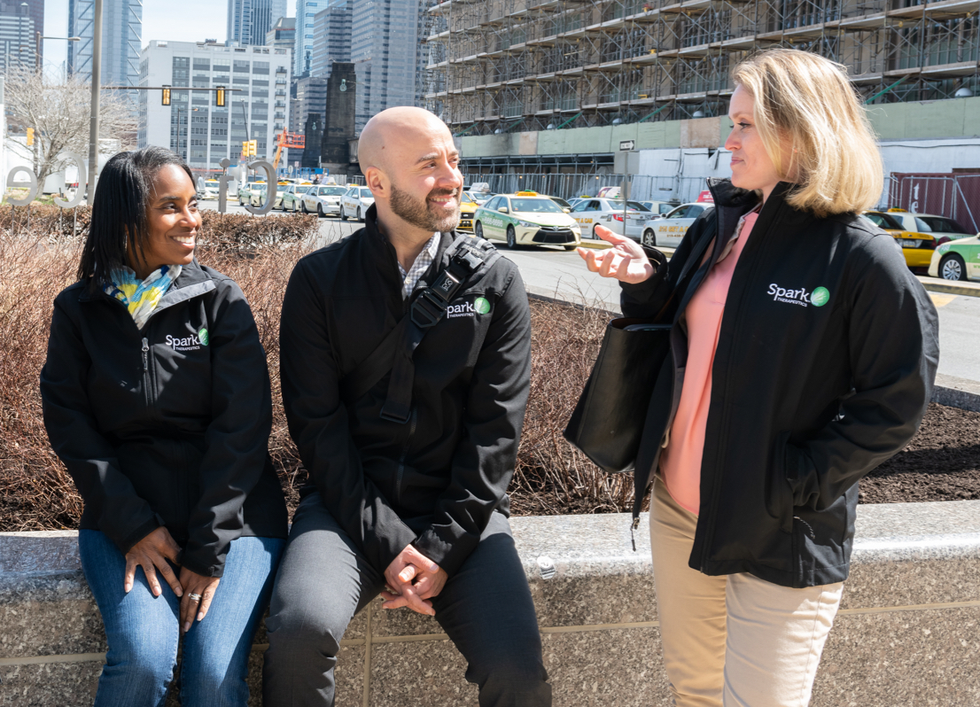 Group of four Spark Therapeutics Medical Affairs team members outdoors with center two seated on a low wall and outer two standing, looking at one team member and smiling with city skyline in background